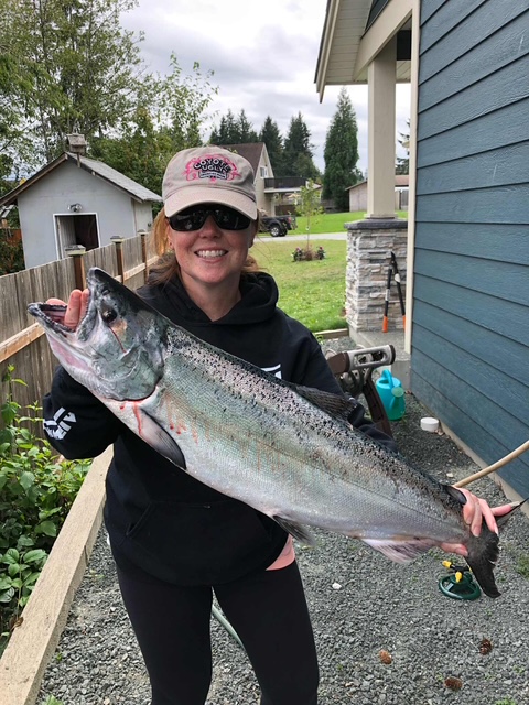 A woman smiling while holding a large fish next to a house on a cloudy day. Gravel, a fence, and a lawn are visible in the background.